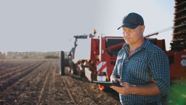 디지털 태블릿을 사용하는 농부. 농사를 짓고 있습니다. 농장 기계들 이 뒤로 처져 있죠. 감자 수확기를 가진 트랙터. 영리 한 농업. 농업 기술. 감자 수확. — 비디오