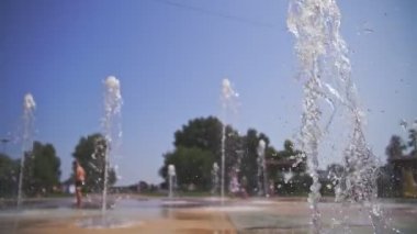Çeşme. Yakın plan. Fıskiye fıskiyeleri. Arka planda bulanık çocuklar, su akıntıları arasında eğleniyorlar, şehir parkında yüksek çeşme jetleri, sıcak yaz günü.