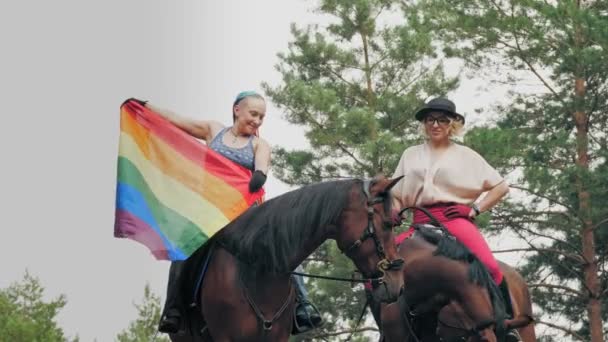 Regnbågsflagga. Kärlek av samma kön. unga, lesbiska, samkönade par ägnar sig åt gemensam hobby, ridning. glada kvinnor som har roligt, hålla regnbåge flagga fladdrar i vinden. — Stockvideo