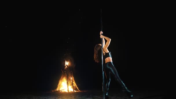 Baile pole. Bruja Halloween. gimnasta femenina en forma, en traje de cuero negro y tacones altos, realiza ejercicios acrobáticos en poste giratorio de metal, por la noche, a la luz de un gran fuego en el fondo. — Vídeos de Stock