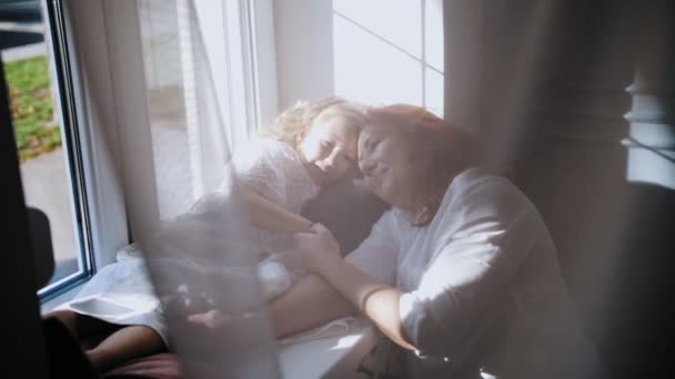 Семья. Дочь мамы. Милая маленькая девочка. Мать и маленькая дочь проводят время вместе, сидя, обнимаясь у окна. — стоковое видео