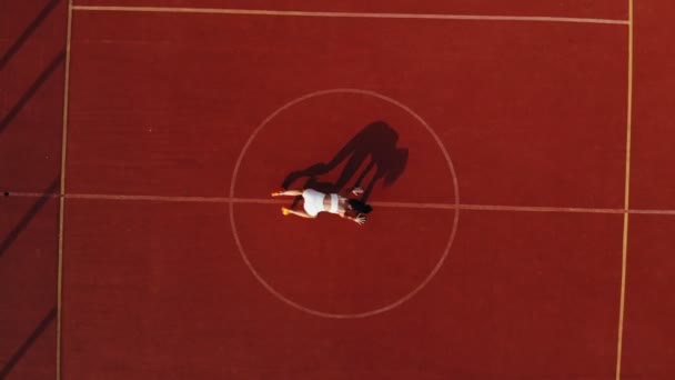 Fitness venku. Aero, pohled shora. Sportovní žena v bílém tričku a šortkách, dělá různá cvičení. pozadí oranžového kurtu s bílými znaky. Venkovní sporty. letní den. na stadionu — Stock video