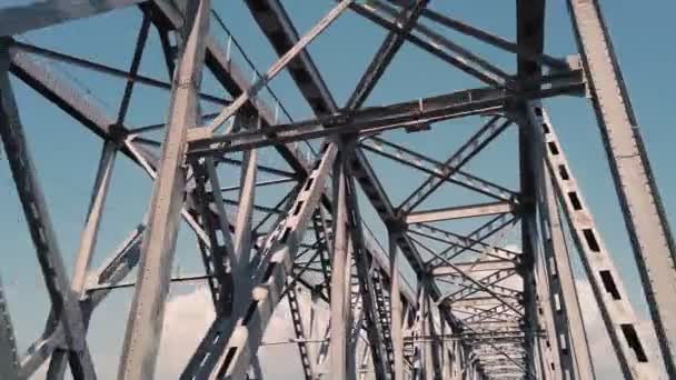 Metallbro. grov järn konstruktioner mot blå himmel, filmad i rörelse från en bil. Bottenvy. Järnvägsbro och vägbro över floden. Silhuett av tvärgående stålbalkar. — Stockvideo