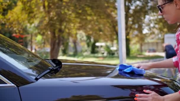 Autowasser. auto vegen. jonge vrouw veegt een auto af met een doek buiten, na het wassen, bij een autowasserette. Auto schoonmaken. — Stockvideo