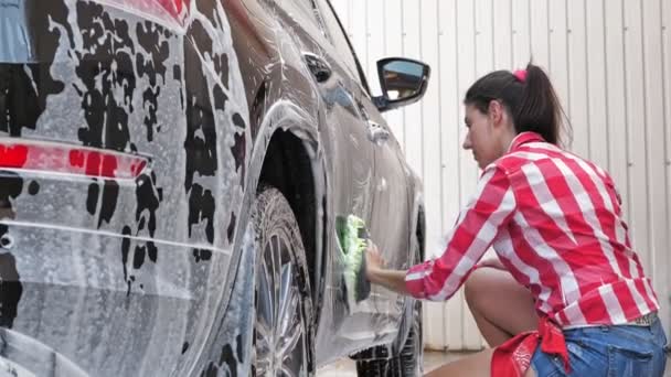 Автомийка. молода жінка, в шортах і сорочці, миє машину, вкрита мильною піною, з мочалкою, самообслуговуванням автомийкою, на відкритому повітрі. Прибирання автомобілів — стокове відео