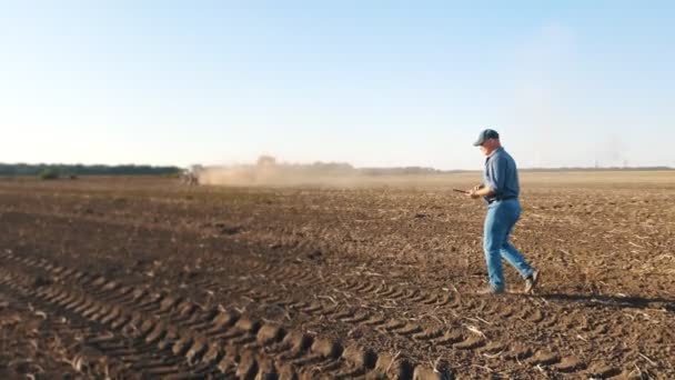 Colheita de batata. agricultor, segurando um tablet digital em suas mãos, caminha através de um campo recém-lavrado contra o pano de fundo de máquinas agrícolas de trabalho, colheitadeiras de batata — Vídeo de Stock