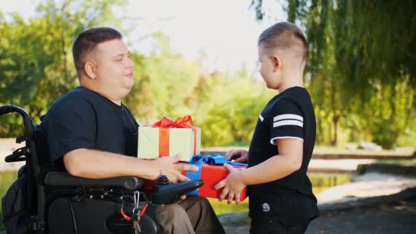 Dzień ojców. Mały, słodki chłopiec daje prezenty swojemu tacie. mężczyzna jest osobą niepełnosprawną. użytkownik wózka inwalidzkiego. osoby o szczególnych potrzebach. — Wideo stockowe