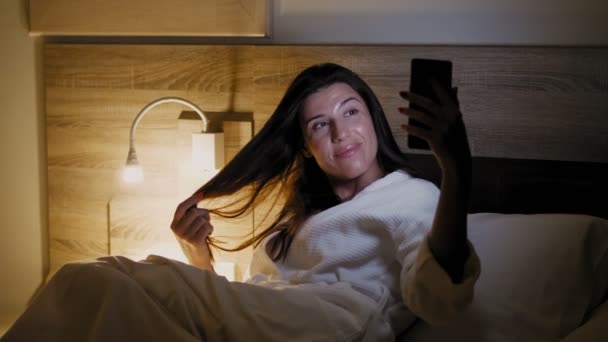 Antes de dormir. uma jovem mulher em um roupão de banho branco fica na cama e usa um smartphone. ao lado da cama, uma luz noturna ou um candeeiro de cabeceira está ligado. tarde da noite ou noite — Vídeo de Stock