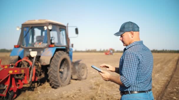 Фермер використовує цифровий планшет. на сільськогосподарському полі, біля трактора, на фоні сільськогосподарської техніки. розумне сільське господарство. технології вирощування — стокове відео