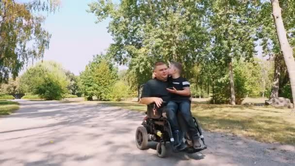 Día de los padres. persona con una discapacidad. Papá y su pequeño hijo lindo están caminando en el parque, en el día de verano. Papá es un usuario de silla de ruedas. personas con necesidades especiales. — Vídeo de stock