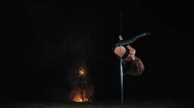Direk dansı. Cadılar Bayramı cadısı. uygun kadın jimnastikçi, siyah deri kıyafetli ve yüksek topuklu, metal döner direkte akrobatik egzersizler yapıyor, geceleri, arka plandaki büyük ateş ışığında,.