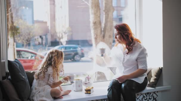 Uma festa de chá. mãe e filha estão bebendo chá com doces, sentados à janela, em um grande peitoril da janela. — Vídeo de Stock