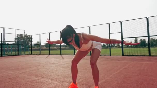 Fitness utomhus. Sportig kvinna, i vit topp och shorts, orange sneakers och solglasögon, gör olika övningar på orange bana med vita markeringar, på stadion. Sommardag. Friluftsliv — Stockvideo