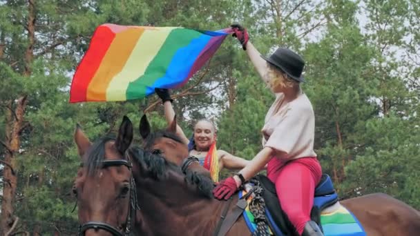 Szivárvány zászló. azonos nemű szerelem. fiatal, leszbikus, azonos nemű pár közös hobbival, lovaglással foglalkozik. boldog nők szórakozás, gazdaság szivárvány zászló lengő szél. — Stock videók