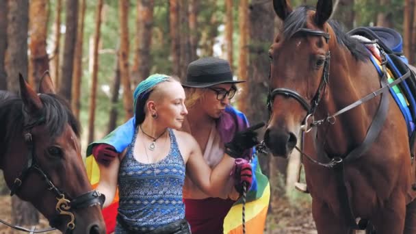 Igbt. Duhová vlajka. láska stejného pohlaví. Mladý lesbický pár stejného pohlaví se angažuje ve společném koníčku, v jízdě na koni. ženy objímající, bavící se, vedle svých koní, v lese — Stock video