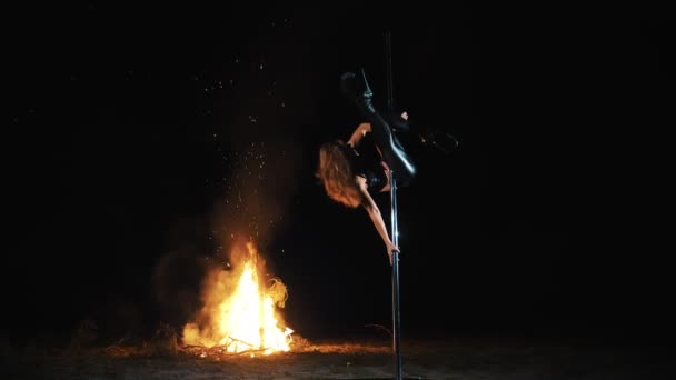 Baile pole. Bruja Halloween. gimnasta femenina en forma, en traje de cuero negro y tacones altos, realiza ejercicios acrobáticos en poste giratorio de metal, por la noche, a la luz de un gran fuego en el fondo. — Vídeos de Stock