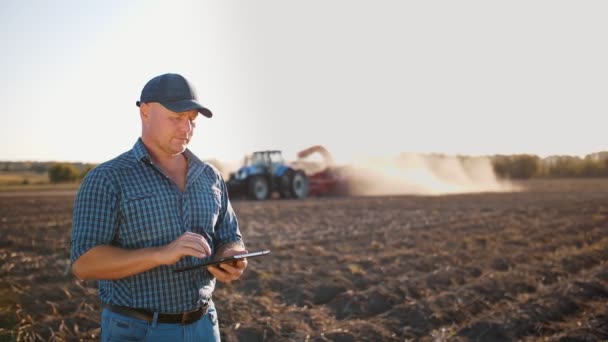 Zbiorów ziemniaków. rolnik używający tabletu cyfrowego. na polu rolniczym, na tle maszyn rolniczych. traktor z kombajnem ziemniaczanym. inteligentne rolnictwo. technologie rolnicze — Wideo stockowe