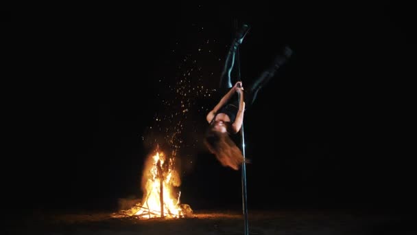 Pole dance. Hexenhalloween. Fitte Turnerin, in schwarzem Lederoutfit und High Heels, führt akrobatische Übungen auf einer rotierenden Stange aus Metall aus, nachts, im Licht des großen Feuers im Hintergrund. — Stockvideo
