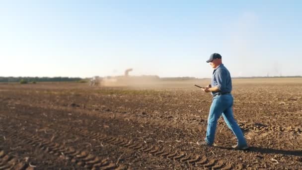 Cosecha de patatas. agricultor, sosteniendo una tableta digital en sus manos, camina a través de un campo recién arado contra el telón de fondo de la maquinaria agrícola de trabajo, cosechadoras de patatas — Vídeo de stock