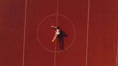 Spor salonu açık havada. Aero, üst manzara. Beyaz bluzlu ve şortlu sportif kadın çeşitli egzersizler yapıyor. Beyaz lekeli turuncu kortun arka planı. Açık hava sporları. Yaz günü. Stadyumda.