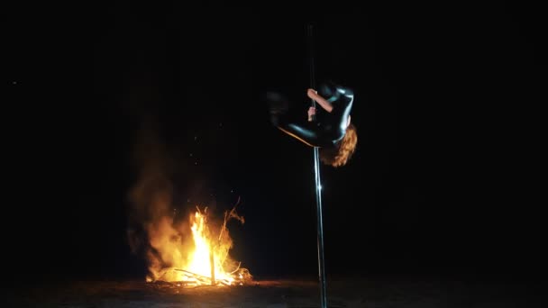 Dança pólo. Bruxa do Dia das Bruxas. caber ginasta feminina, em roupa de couro preto e salto alto, realiza exercícios acrobáticos no poste giratório de metal, à noite, pela luz do grande fogo no fundo. — Vídeo de Stock