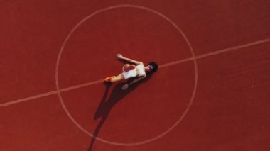 Spor salonu açık havada. Aero, üst manzara. Beyaz bluzlu ve şortlu sportif kadın çeşitli egzersizler yapıyor. Beyaz lekeli turuncu kortun arka planı. Açık hava sporları. Yaz günü. Stadyumda.