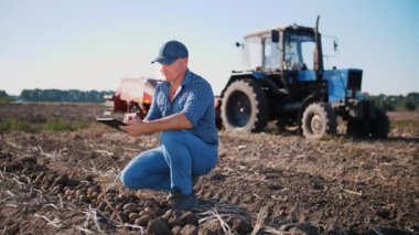 Patates hasadı. Dijital tablet kullanan çiftçi. Çiftlik arazisinde, çiftlik makinelerinin arka planında. Patates biçme makineli traktör. Akıllı çiftçilik. tarım teknolojileri