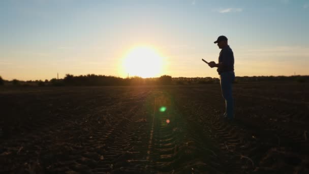Földműves sziluett. A farmer egy digitális táblán dolgozik. szabadban, mezőgazdasági területen, naplementekor. a háttérvilágításban. gazdálkodás — Stock videók