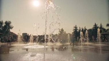 Çeşme. Yakın plan. Fıskiye fıskiyeleri. Arka planda bulanık çocuklar, su akıntıları arasında eğleniyorlar, şehir parkında yüksek çeşme jetleri, sıcak yaz günü.