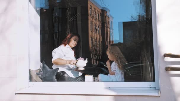 Uma festa de chá. mãe e filha estão bebendo chá com doces, biscoitos, sentados à janela, em um grande peitoril da janela. vista através da janela, ao ar livre — Vídeo de Stock