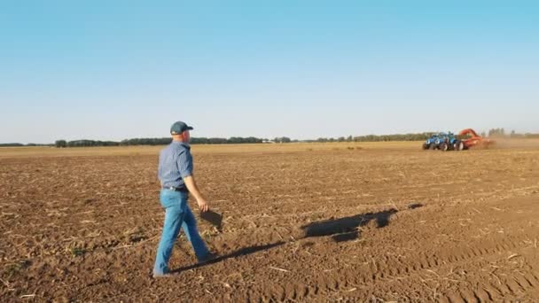 Cosecha de patatas. agricultor, sosteniendo una tableta digital en sus manos, camina a través de un campo recién arado contra el telón de fondo de la maquinaria agrícola de trabajo, cosechadoras de patatas — Vídeos de Stock