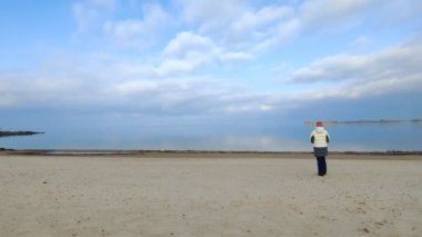 landscape of deserted beach. a teenage girl walks along sand shore near the river, lake or sea, in cold weather. early spring. blue sky with white clouds.