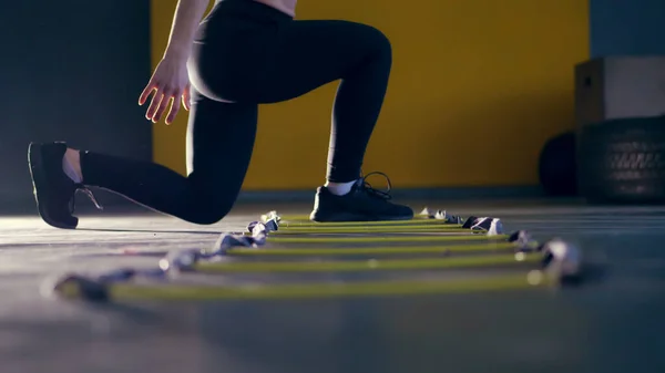 close-up, female legs in black leggings and sneakers. woman performs exercises with an athletes foot ladder, jumps , training on agility ladder on floor, in gym. Fitness class cardio workout .