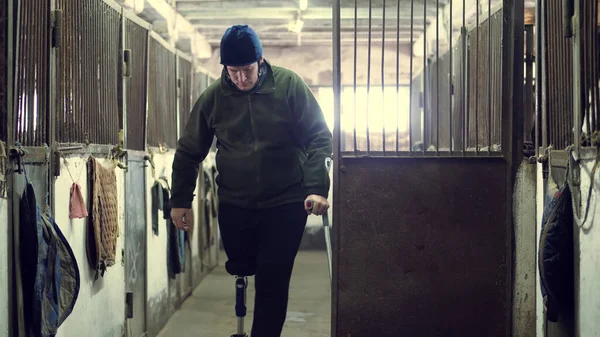 in stable, man with disability puts on helmet for riding, he walks, leaning on crutch, through stable. man has artificial limb instead of his right leg. concept of rehabilitation of the disabled with