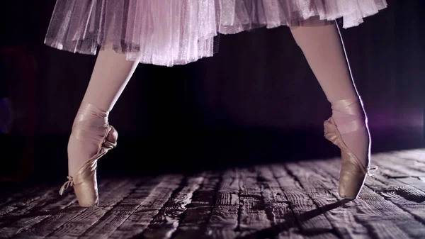 close up, in rays of spotlight, on the stage of the old theater hall. ballerina in white ballet skirt, raises on toes in pointe shoes, performs elegantly a certain ballet exercise, relleve. High