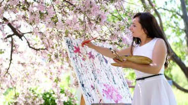a beautiful woman painter in white dress, artist paints a picture of flowers in blooming spring apple orchard, she holds a palette with paints and a brush in her hands. High quality photo