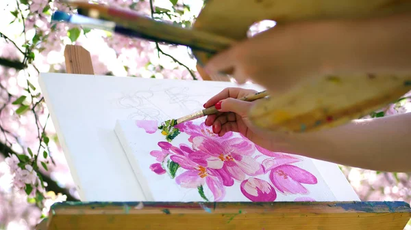 close-up, painter draw a picture of flowers in blooming spring apple orchard, she paints with brush, holds palette with paints. wind sweeps flowering branches over the easel. High quality photo