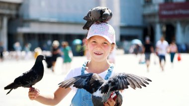 VENICE, ITALY - JULY 7, 2018: view of happy kid girl, tourist, holding pigeons, feeding, play with them, having fun on Piazza San Marco, St Marks Basilica, on a summer day. High quality photo