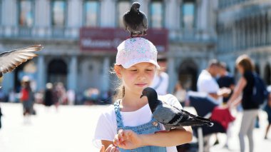 VENICE, ITALY - JULY 7, 2018: view of happy kid girl, tourist, holding pigeons, feeding, play with them, having fun on Piazza San Marco, St Marks Basilica, on a summer day. High quality photo