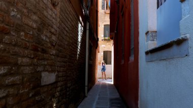 VENICE, ITALY - JULY 7, 2018: at the end of a very narrow street in Venice, a young woman tourist, with a hat and a backpack, stands and looks at, examines architecture. High quality photo