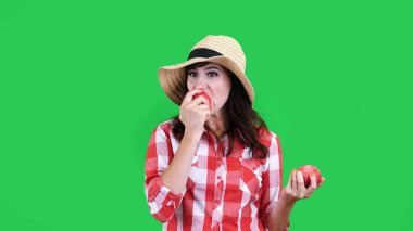 Komik, kareli gömlekli ve şapkalı kadın çiftçi, iki büyük ekşi elmayı tutuyor, yiyor, ısırıyor, yüzünü kırıştırıyor, stüdyoda yeşil arka planda. Masanız için sağlıklı yemek. Yüksek