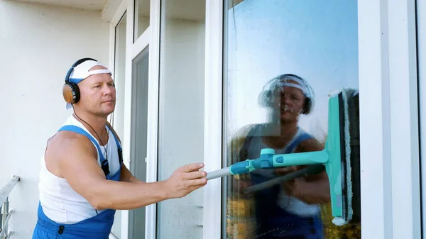 白人有魅力的男人 清洁服务人员 穿着蓝色工作服 用耳机听手机上的音乐 用特殊的拖把清洁 用特殊的拖把洗窗 高质量的照片 — 图库照片