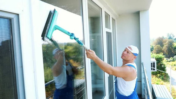 白人英俊的男人 清洁服务的工人 穿着蓝色工作服 头戴帽子 漂亮剂 用特殊的拖把清洗窗户 享受他的工作 高质量的照片 — 图库照片