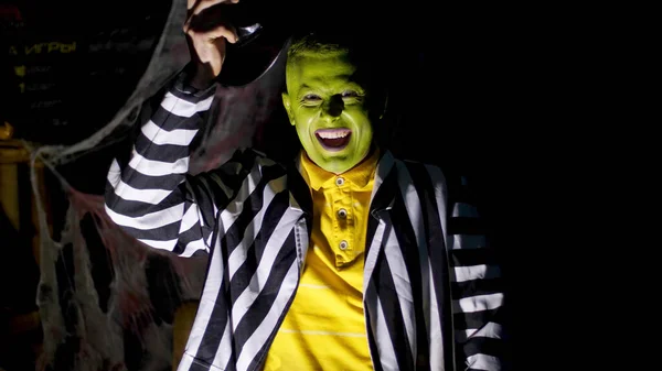 Halloween party, night, twilight, in the rays of light, a man with a terrible make-up, with a green face lifts his hat, laughs eerily and runs away. Behind the background is a spiderweb. High quality