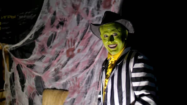 Halloween party, night, twilight, in the rays of light, a man with a terrible make-up, with a green face and a hat shows fear and runs away. Behind the background is a spiderweb. High quality photo