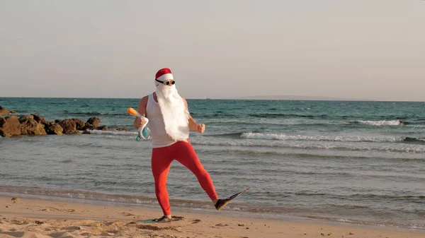 Santa Claus summer vacation. Santa Claus having fun. Funny Santa, in sunglasses, with flippers and snorkeling mask, is dancing on beach by the sea. Santa going to snorkel. High quality photo