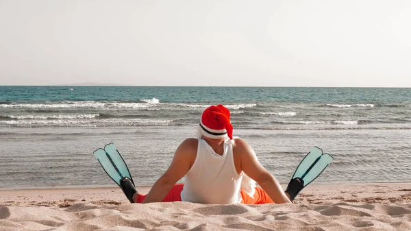 Santa Claus summer vacation. Santa Claus having fun. Funny Santa, in flippers, relaxing while sitting on sandy beach by the sea. back view. High quality photo
