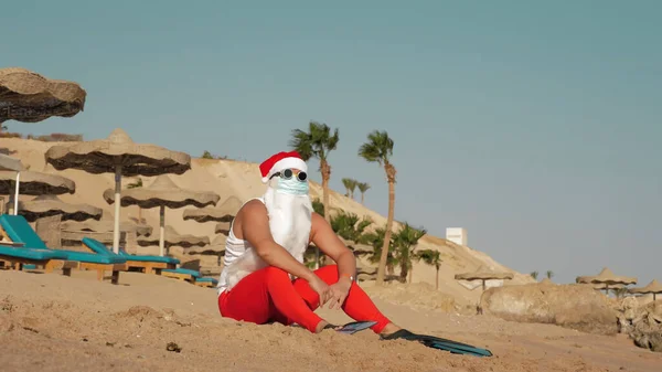 Santa Claus summer vacation. Santa Claus having fun. Funny Santa, in protective mask, sunglasses and flippers, relaxing while lying on sandy beach by the sea, among palm trees. High quality photo