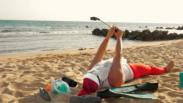 Santa Claus blogger. Santa Claus takes selfie. Funny Santa, in sunglasses, takes selfie video, while lying on sandy beach by the sea. Santa summer vacation. High quality photo