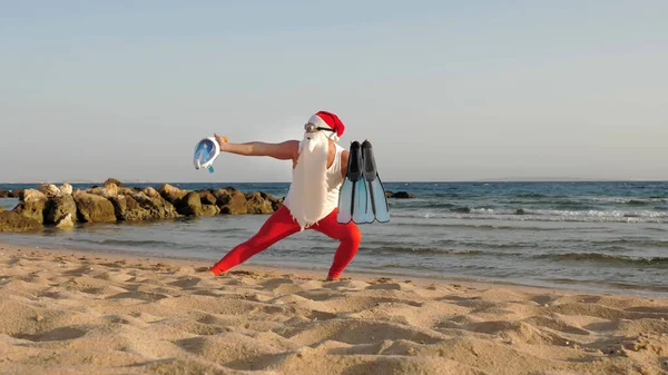 Santa Claus summer vacation. Father Christmas, having fun. Santa Claus doing yoga. Funny Santa, in sunglasses, with flippers and snorkeling mask, on beach by the sea. Santa going to snorkel. High